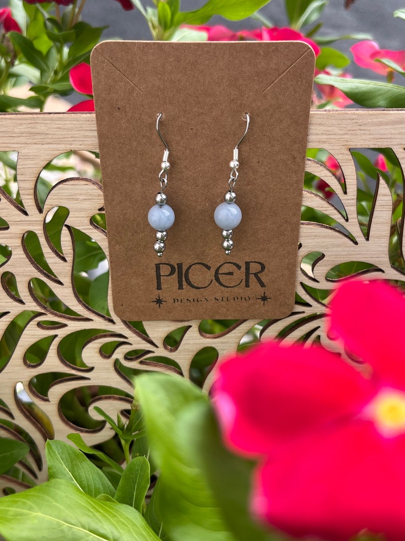 May include: A pair of silver earrings with light blue round gemstone beads. The earrings are displayed on a brown card with the text "Picer Design Studio".