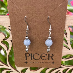 May include: A pair of silver earrings with light blue gemstone beads. The earrings are on a brown card with the text "Picer Design Studio".