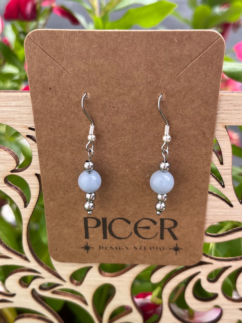 May include: A pair of silver earrings with light blue gemstone beads. The earrings are on a brown card with the text "Picer Design Studio".