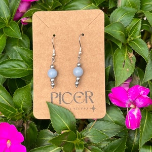 May include: A pair of silver dangle earrings with light blue round beads. The earrings are displayed on a brown card with the text "Picer Studio" printed on it.