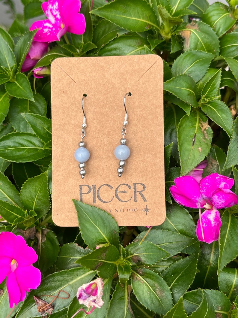 May include: A pair of silver dangle earrings with light blue round beads. The earrings are displayed on a brown card with the text "Picer Studio" printed on it.
