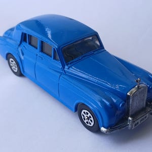 Könnte beinhalten: Ein leuchtend blaues Spielzeugauto aus Druckguss mit silberfarbenem Kühlergrill, schwarzen Rädern mit silbernen Speichen und einem Nummernschild an der vorderen Stoßstange mit der Aufschrift RR 1959. Das Fahrzeug hat eine klassische Limousinenform.