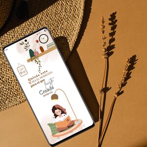 Puede incluir: Un smartphone muestra un fondo de pantalla digital con una mujer leyendo un libro bajo una lámpara. El fondo de pantalla incluye texto en español, una estantería con jarrones y un reloj, y un icono en forma de corazón. El teléfono está sobre una superficie marrón.