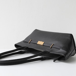 May include: Black leather tote bag with a rectangular shape. The bag features a gold-colored rectangular clasp and two gold-colored bars. The bag has two black handles and a zipper closure.