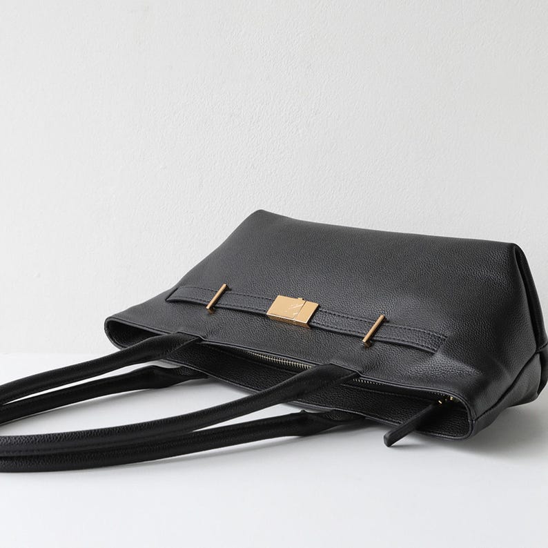 May include: Black leather tote bag with a rectangular shape. The bag features a gold-colored rectangular clasp and two gold-colored bars. The bag has two black handles and a zipper closure.