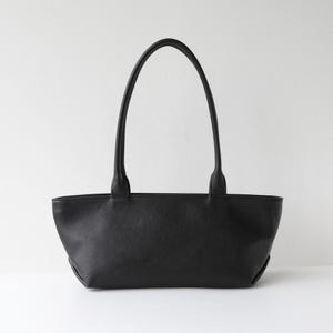 May include: A black tote bag with two long handles. The bag is made of a textured material and has a rectangular shape with rounded corners. The handles are attached to the top of the bag and are long enough to be carried over the shoulder.