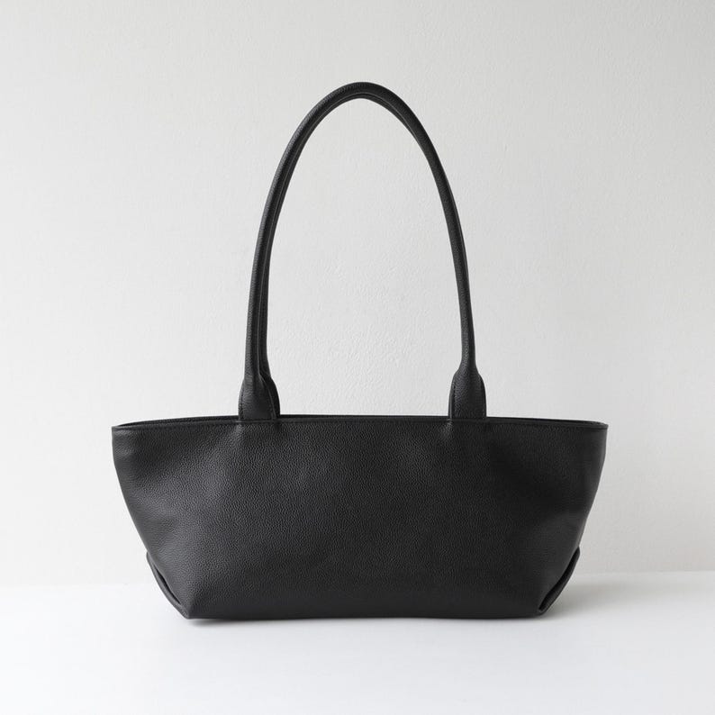 May include: A black tote bag with two long handles. The bag is made of a textured material and has a rectangular shape with rounded corners. The handles are attached to the top of the bag and are long enough to be carried over the shoulder.