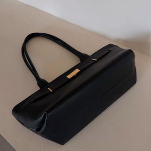 May include: A black leather tote bag with a rectangular shape and two handles. The bag features gold-colored accents on the front and a textured surface. The bag is designed for carrying items.