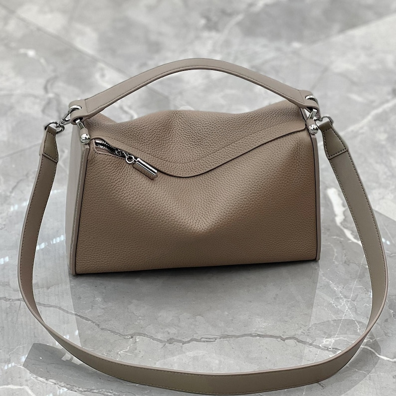 May include: A taupe-colored handbag with a top handle and a long, adjustable shoulder strap. The bag features a textured leather exterior, a silver zipper, and silver hardware. The design is a rectangular shape with a curved top.