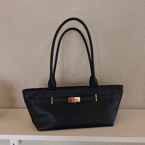 May include: Black tote bag with two long handles. The bag has a rectangular shape with a gold-colored clasp and accents. The bag is sitting on a white shelf.