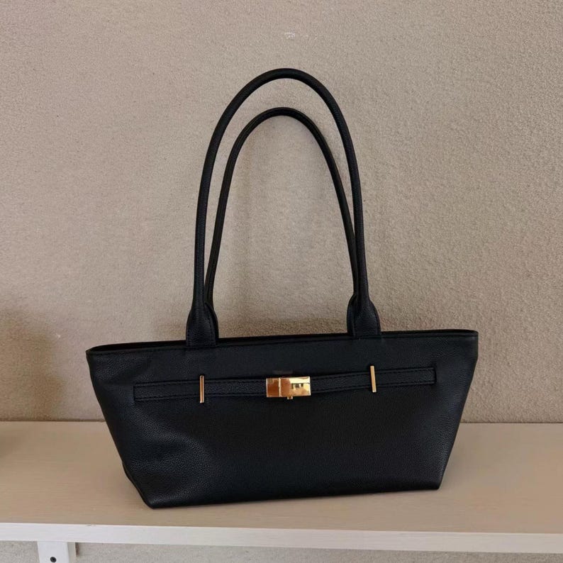 May include: Black tote bag with two long handles. The bag has a rectangular shape with a gold-colored clasp and accents. The bag is sitting on a white shelf.