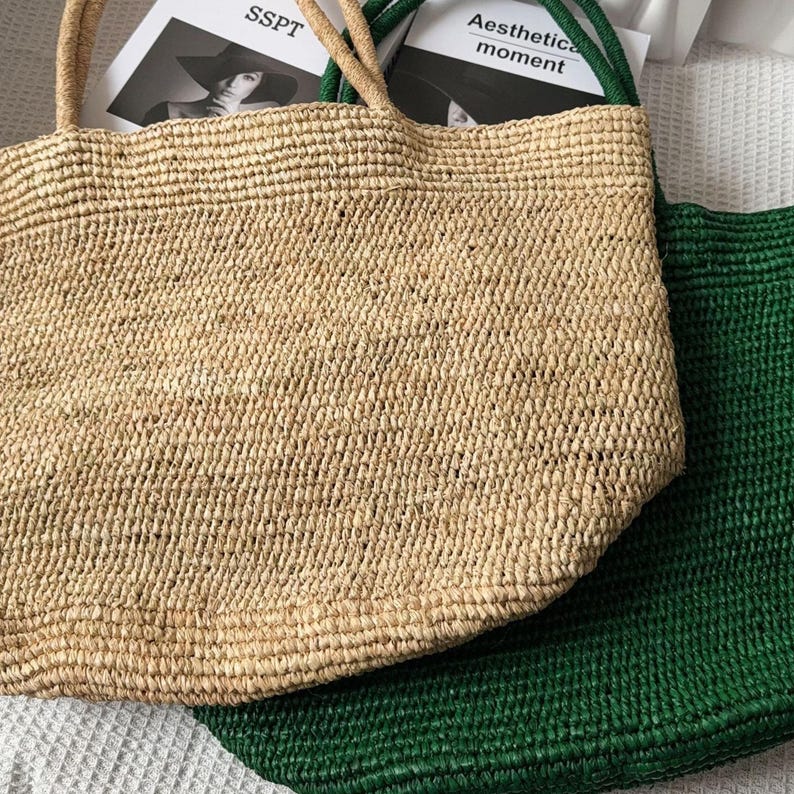 May include: A tan woven straw tote bag with two matching handles positioned in front of a dark green woven straw tote bag. The tan bag features a rectangular body with a textured, braided surface and two top handles for carrying by hand or shoulder.