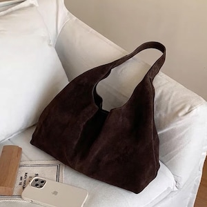 May include: A dark brown suede tote bag with a wide, open design and long handles. The bag is resting on a white surface. The bag's color is a rich, earthy tone, and the material appears soft and textured.
