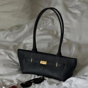 May include: Black handbag with a rectangular shape and two long handles. The bag features a gold-colored clasp and accents. The bag is sitting on a white surface, with sunglasses and a small container visible in the foreground.