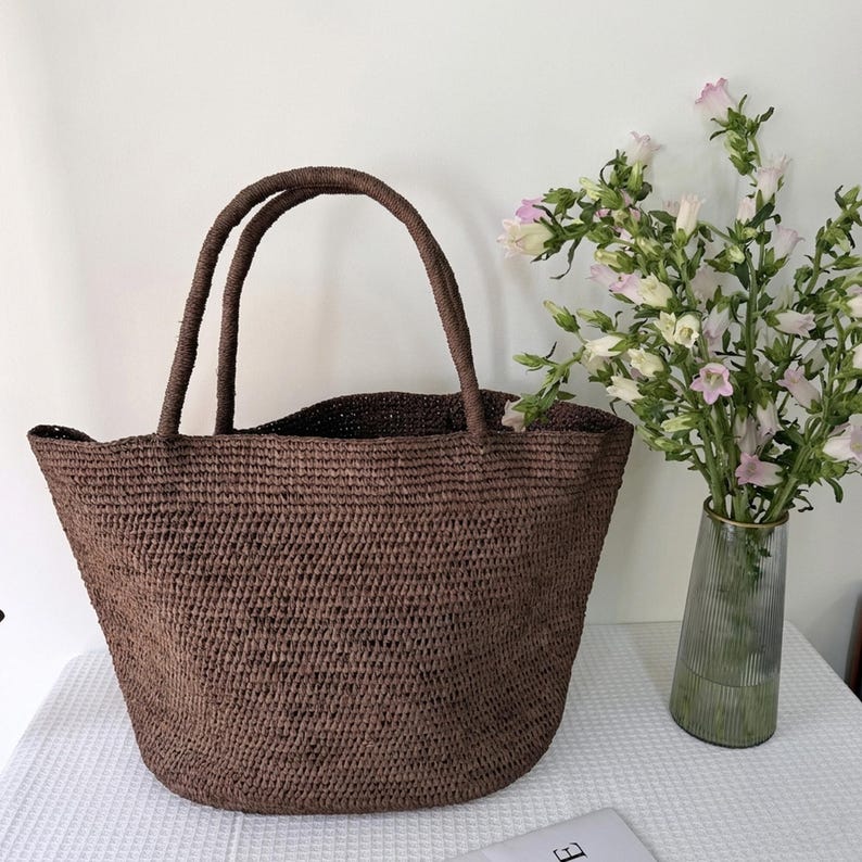 May include: A dark brown woven tote bag with two matching handles, featuring a textured, open-weave construction. The bag has a wide, rounded base and a spacious, open top design, providing a simple and functional structure for carrying items.