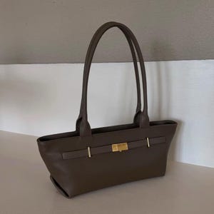 May include: A brown leather tote bag with two long handles. The bag features a rectangular shape with a gold-colored clasp and accents. The bag is sitting on a white surface.