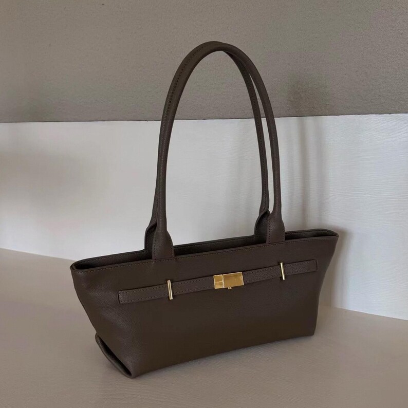 May include: A brown leather tote bag with two long handles. The bag features a rectangular shape with a gold-colored clasp and accents. The bag is sitting on a white surface.