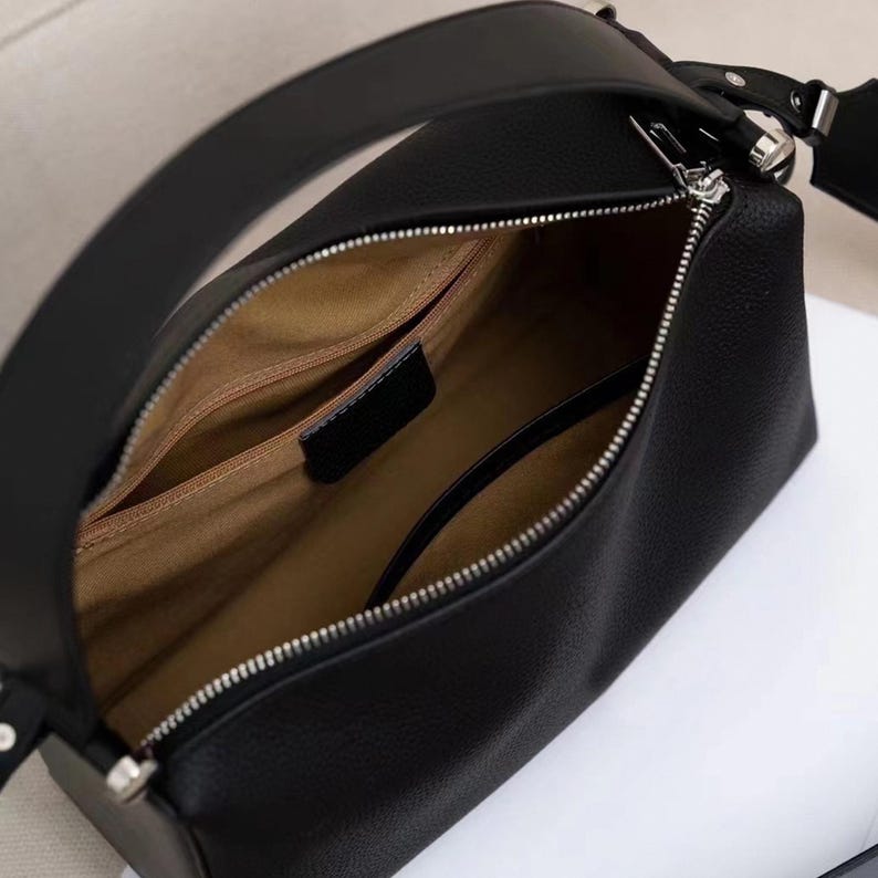May include: Black leather handbag with a silver zipper and a long shoulder strap. The interior is tan with multiple compartments. The bag has a rectangular shape with rounded edges.