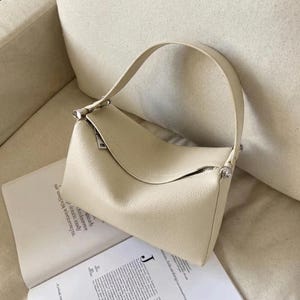 May include: Cream-colored handbag with a curved flap and a single handle. The bag has a rectangular shape with a zipper closure and silver-toned hardware. The bag is sitting on top of an open book.