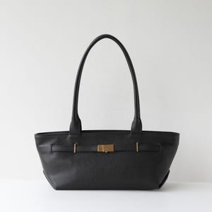 May include: Black leather tote bag with a rectangular shape and two long handles. The bag features a gold-colored buckle closure and a strap detail across the front. The bag is set against a white background.