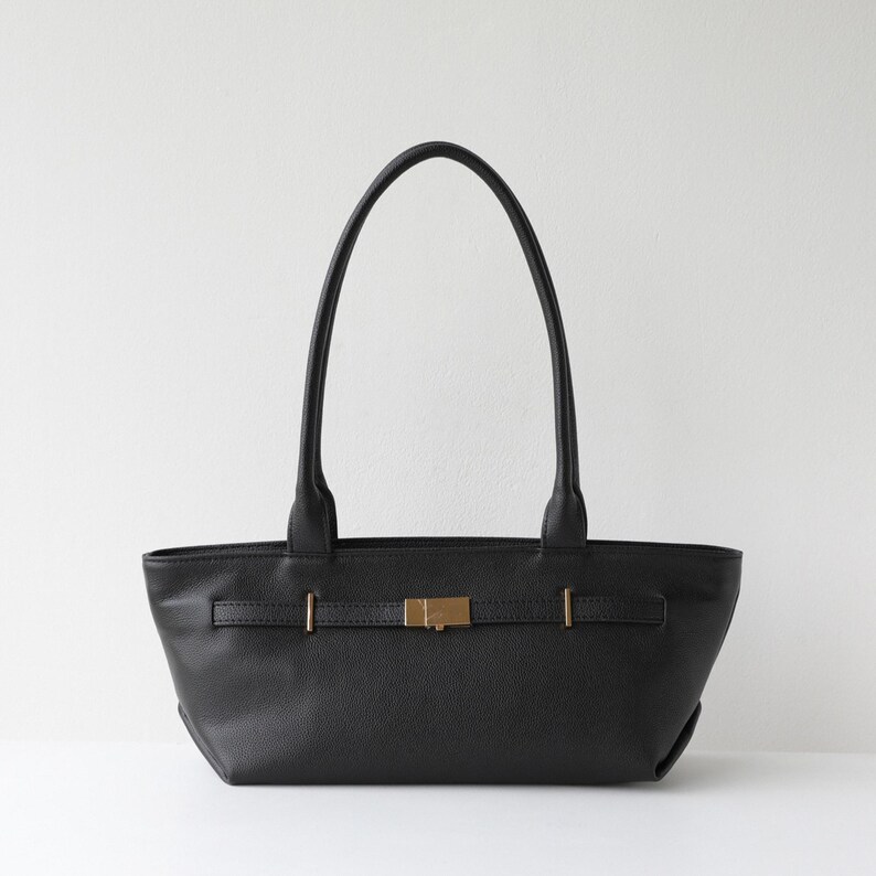 May include: Black leather tote bag with a rectangular shape and two long handles. The bag features a gold-colored buckle closure and a strap detail across the front. The bag is set against a white background.