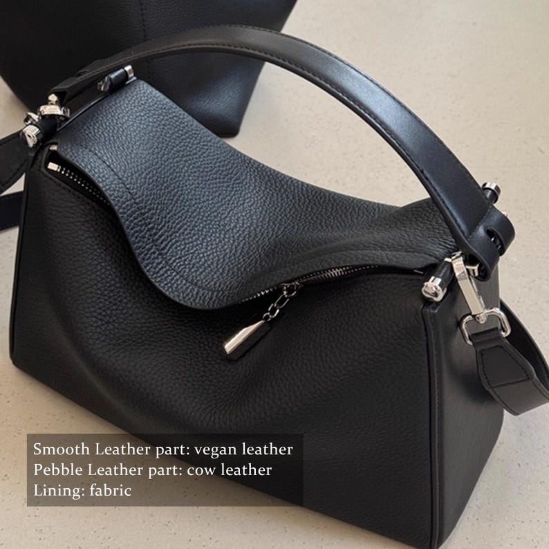 May include: Black handbag with a curved top handle and a detachable shoulder strap. The bag features a zipper closure with a silver-toned pull. The smooth leather part is vegan leather, and the pebble leather part is cow leather. The lining is fabric.