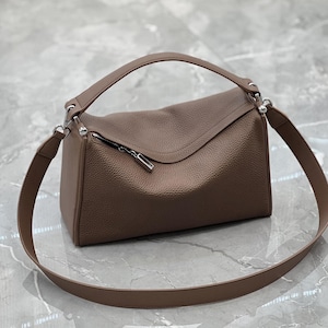May include: A brown leather handbag with a top handle and a long shoulder strap. The bag has a rectangular shape with a curved flap and a silver zipper. The leather has a textured appearance.