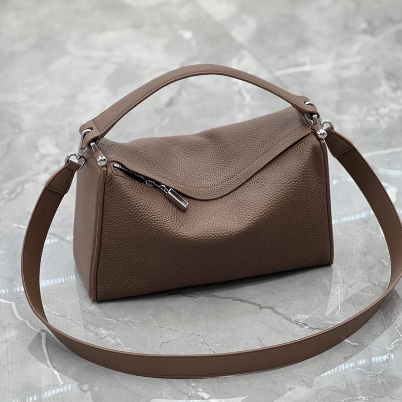 May include: A brown leather handbag with a top handle and a long shoulder strap. The bag has a rectangular shape with a curved flap and a silver zipper. The leather has a textured appearance.