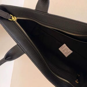 May include: Black leather tote bag with gold-tone zipper closures and handles. The interior features a large compartment and a smaller zippered pocket. A small white tag with text is visible inside the bag.