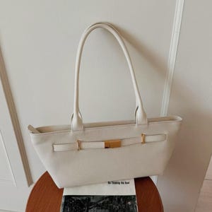 May include: Cream-colored tote bag with long handles and a rectangular shape. The bag features a gold-colored clasp and buckle detail on the front. The bag is sitting on a wooden table.