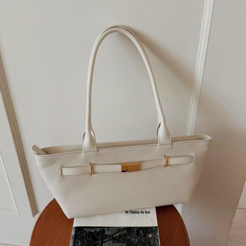 May include: Cream-colored tote bag with long handles and a rectangular shape. The bag features a gold-colored clasp and buckle detail on the front. The bag is sitting on a wooden table.