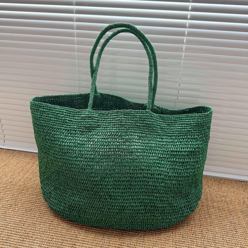 May include: A deep forest green tote bag crafted from woven straw material. The bag features a wide, open-top design with two matching woven handles attached to the top rim. The texture is consistent throughout the body, showing a dense, horizontal weave pattern that creates a sturdy, structured shape for this everyday carry accessory.