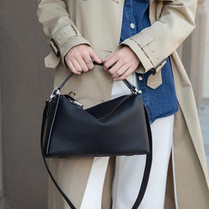 Könnte beinhalten: Schwarze Lederhandtasche mit strukturiertem Design. Die Tasche hat einen oberen Griff und einen abnehmbaren Schultergurt. Die Tasche wird von einer Person gehalten, die einen beigen Trenchcoat, ein Jeanshemd und eine weiße Hose trägt.