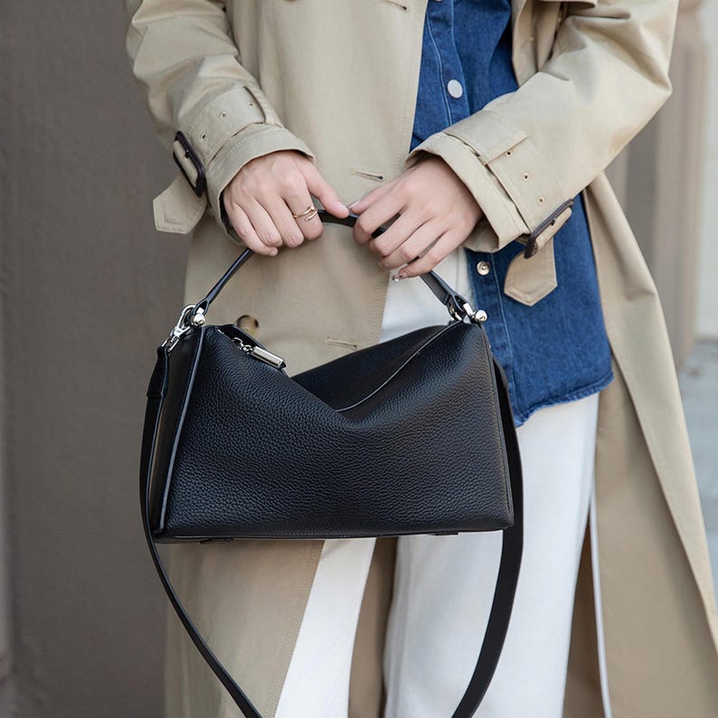 May include: Black leather handbag with a structured design. The bag features a top handle and a detachable shoulder strap. The bag is held by a person wearing a tan trench coat, a denim shirt, and white pants.