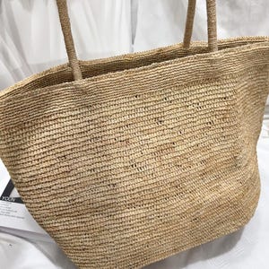 May include: A large, tan-colored tote bag constructed from woven natural fibers. The bag features a wide, open-top design with two sturdy, matching woven handles attached to the upper rim. The texture is consistent throughout the body.
