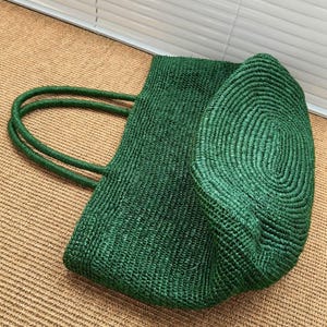 May include: A deep forest green tote bag crafted from woven straw material. The bag features a rectangular body with a rounded, coiled base and two matching green woven handles attached to the top edge for carrying the bag by hand or shoulder.