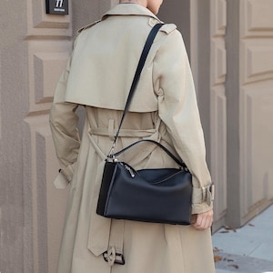 May include: A black leather handbag with a shoulder strap and top handle. The bag has a rectangular shape with a curved top and a silver zipper. The person is wearing a tan trench coat.