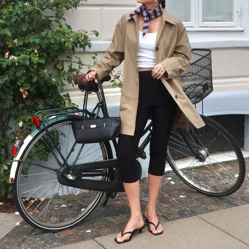 May include: A woman stands next to a green bicycle, wearing a tan trench coat, white top, and black leggings. She has a patterned scarf and black sandals. A black purse hangs on the bicycle. The bicycle has a black basket and a red reflector.