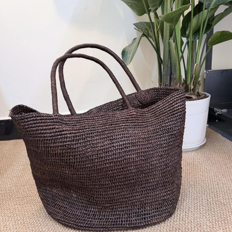 May include: A dark brown woven tote bag featuring a textured, straw-like construction with two rounded handles attached to the top. The bag has a wide, open-top design with a consistent, dense weave pattern throughout the entire exterior surface.