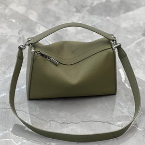 May include: Olive green leather handbag with a top handle and a detachable shoulder strap. The bag features a curved top design and a silver zipper closure. The leather has a textured appearance.