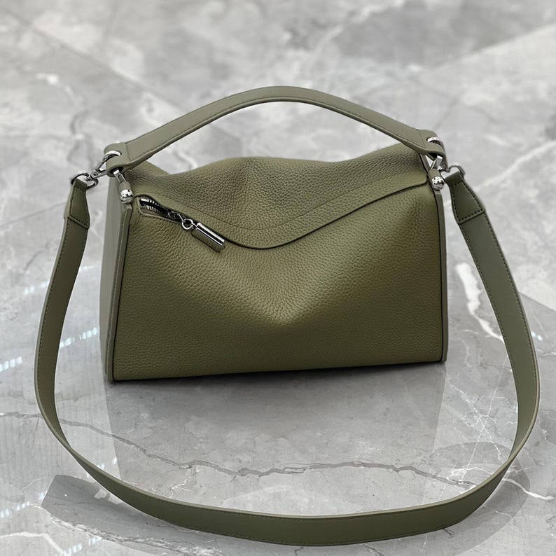 May include: Olive green leather handbag with a top handle and a detachable shoulder strap. The bag features a curved top design and a silver zipper closure. The leather has a textured appearance.