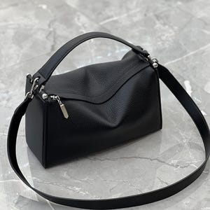 May include: A black leather handbag with a top handle and a detachable shoulder strap. The bag has a rectangular shape with a flap closure and silver-tone hardware. The leather has a textured appearance.