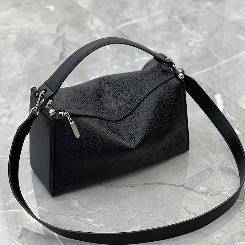 May include: A black leather handbag with a top handle and a detachable shoulder strap. The bag has a rectangular shape with a flap closure and silver-tone hardware. The leather has a textured appearance.