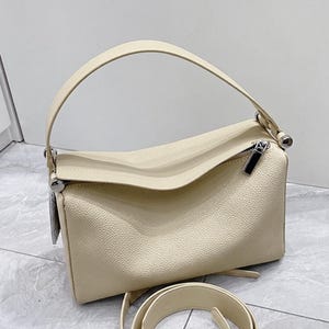 May include: Cream-colored handbag with a top handle and a detachable shoulder strap. The bag features a rectangular shape with a textured surface and a silver zipper. The design is simple and elegant, suitable for everyday use.
