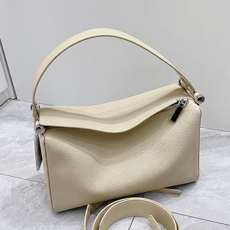 May include: Cream-colored handbag with a top handle and a detachable shoulder strap. The bag features a rectangular shape with a textured surface and a silver zipper. The design is simple and elegant, suitable for everyday use.