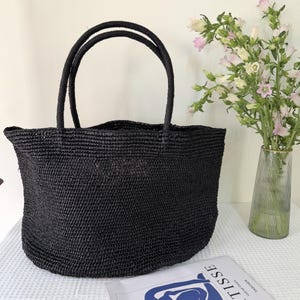 May include: A black woven tote bag with two matching rounded handles. The bag features a textured, straw-like construction with a wide, open-top design. The material has a consistent dark finish throughout the entire body and the handles.