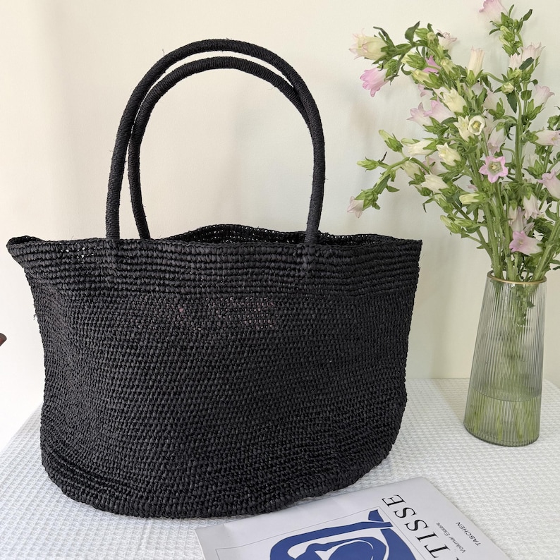 May include: A black woven tote bag with two matching rounded handles. The bag features a textured, straw-like construction with a wide, open-top design. The material has a consistent dark finish throughout the entire body and the handles.