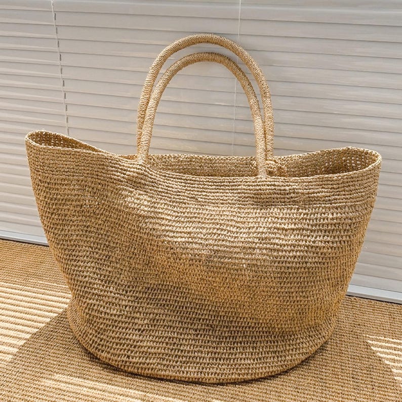 May include: A large, open-top tote bag crafted from natural tan woven straw. The bag features a wide, rounded base that tapers toward the top, two sturdy integrated handles, and a consistent, textured knit pattern throughout the entire structure.