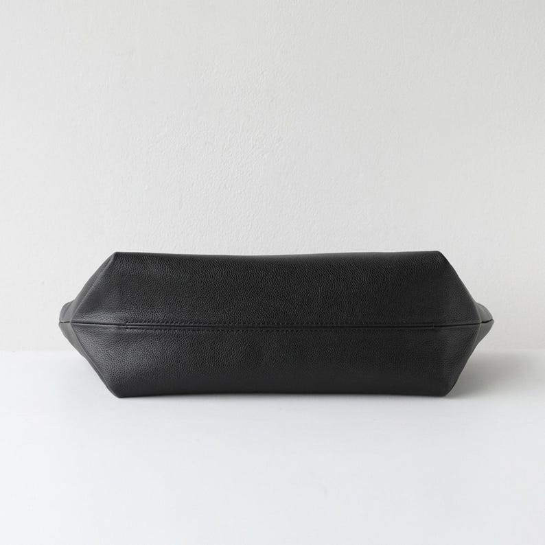 May include: A black leather tote bag with a rectangular shape. The bag has a textured surface and a stitched seam running across the center. The bag is set against a white background.