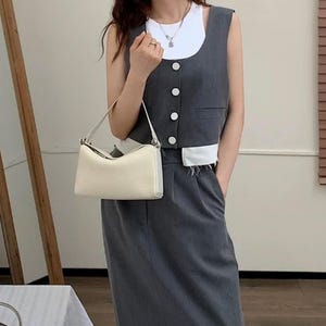 May include: A cream-colored, rectangular handbag with a single shoulder strap. The bag is held in front of a person wearing a gray vest with white trim and a matching skirt. The vest has white buttons and a pocket.